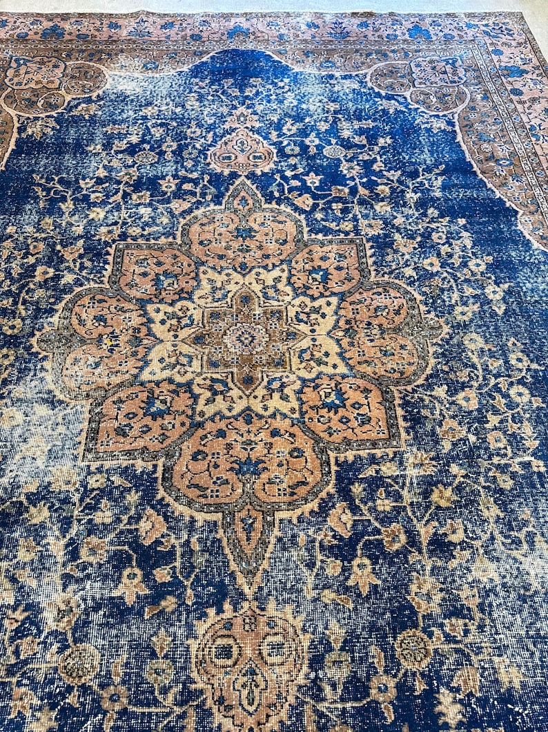 May include: A distressed, vintage-style rug with a central medallion design. The rug features a deep blue field with intricate floral patterns in shades of beige and rust. The edges have a faded appearance, adding to its antique aesthetic.