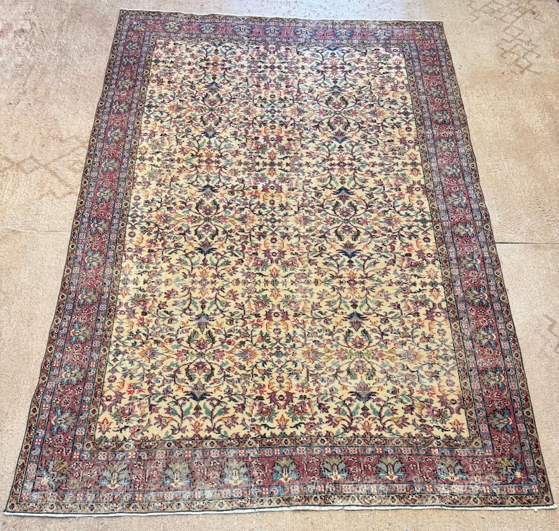 May include: An antique Persian rug with a cream-colored field and an intricate floral pattern in shades of red, blue, green, and gold. The rug has a detailed border in red and cream.