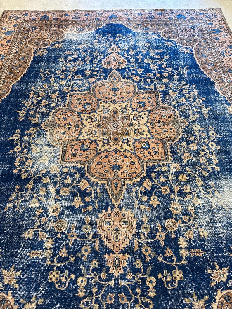 May include: A large, rectangular rug with a distressed, vintage appearance. The rug features a deep blue central field with a central medallion and floral patterns in shades of peach, tan, and brown. The border is a complementary color palette.