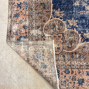 May include: A distressed, vintage-style rug with a folded corner, showcasing a blend of colors. The rug features a navy blue central field with floral patterns, bordered by a brown and blush pink design. The rug's edges have a fringed detail.