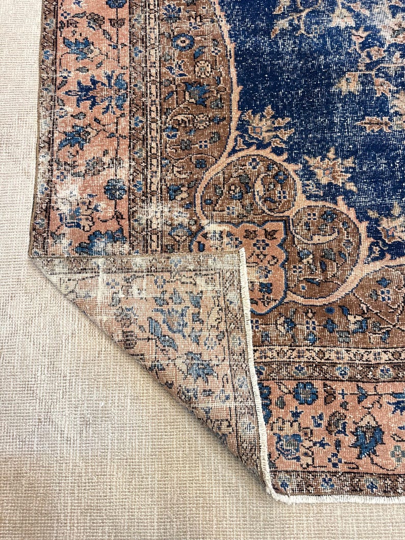 May include: A distressed, vintage-style rug with a folded corner, showcasing a blend of colors. The rug features a navy blue central field with floral patterns, bordered by a brown and blush pink design. The rug's edges have a fringed detail.