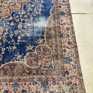May include: A distressed, vintage-style rug with a central field of deep blue, accented by faded areas. The rug features intricate floral and geometric patterns in shades of brown, beige, and light pink. The border is a lighter pink with blue floral motifs.
