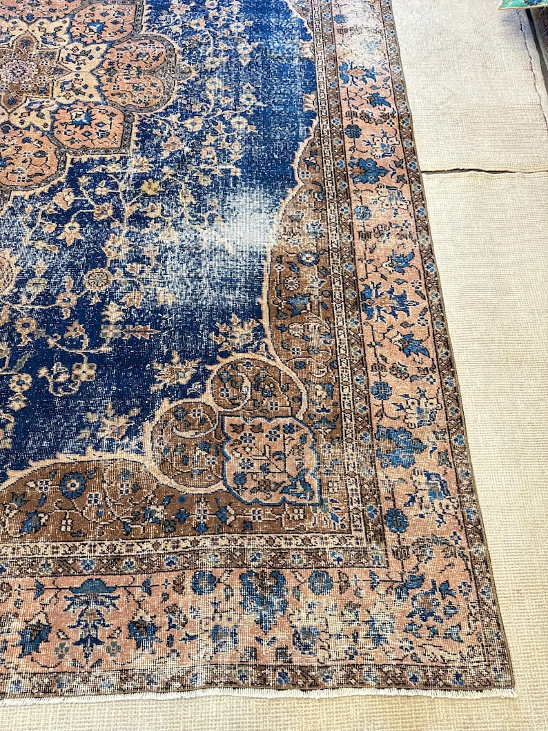 May include: A distressed, vintage-style rug with a central field of deep blue, accented by faded areas. The rug features intricate floral and geometric patterns in shades of brown, beige, and light pink. The border is a lighter pink with blue floral motifs.