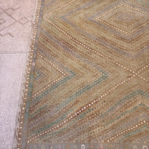 May include: A rectangular rug with a geometric diamond pattern. The rug features a color palette of tan, brown, blue, and white. The edges of the rug have a decorative border and fringe.