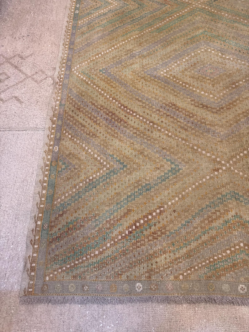 May include: A rectangular rug with a geometric diamond pattern. The rug features a color palette of tan, brown, blue, and white. The edges of the rug have a decorative border and fringe.