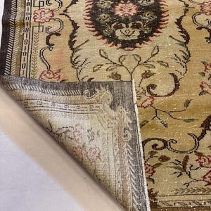 May include: A vintage beige rug with a detailed floral pattern in shades of brown, red, and green. The rug has a distressed look, with a folded corner revealing the underside. The design includes a central medallion and ornate borders.