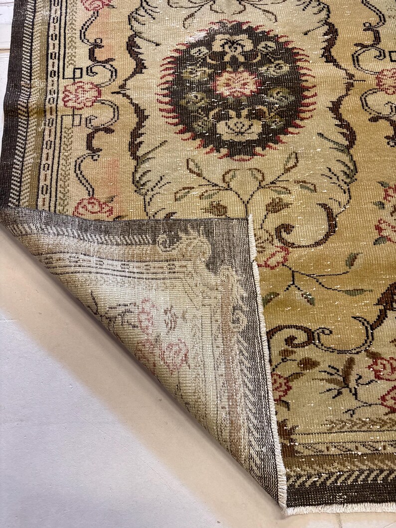 May include: A vintage beige rug with a detailed floral pattern in shades of brown, red, and green. The rug has a distressed look, with a folded corner revealing the underside. The design includes a central medallion and ornate borders.