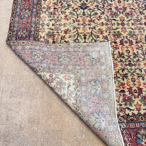 May include: A section of a vintage rug with a cream-colored field and a detailed floral pattern in shades of red, blue, green, and purple. The rug's border is a deep red with intricate designs. The rug is partially folded, revealing its underside.
