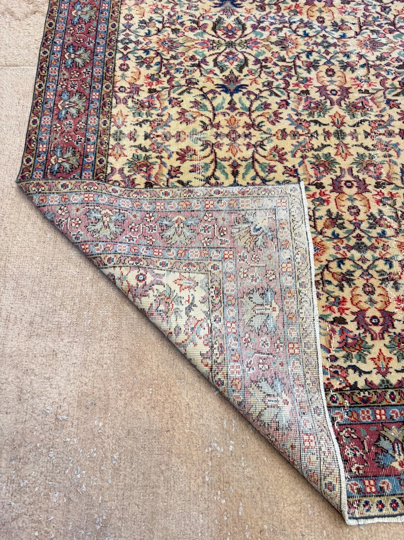 May include: A section of a vintage rug with a cream-colored field and a detailed floral pattern in shades of red, blue, green, and purple. The rug's border is a deep red with intricate designs. The rug is partially folded, revealing its underside.