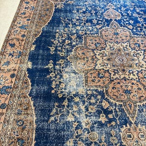 May include: A distressed, vintage-style rug with a central navy blue field and intricate floral patterns in shades of peach, brown, and cream. The rug features a detailed border and a faded, antique appearance, suitable for home decor.