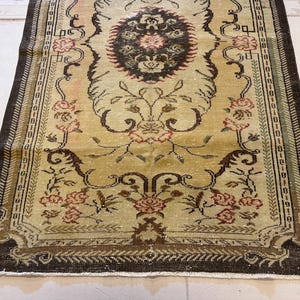 May include: A rectangular, vintage-style rug with a central medallion design. The rug features a cream-colored field with floral and scroll motifs in shades of brown, red, and green. The border is a dark brown with a repeating pattern.