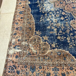 May include: A section of a vintage rug with a distressed appearance. The rug features a deep blue central field with intricate floral patterns in shades of brown and peach. The border is a peach color with blue floral designs.