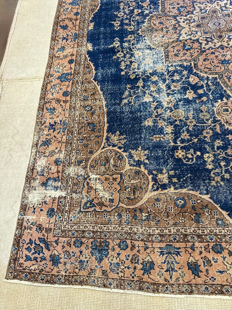 May include: A section of a vintage rug with a distressed appearance. The rug features a deep blue central field with intricate floral patterns in shades of brown and peach. The border is a peach color with blue floral designs.