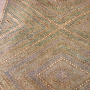 May include: A vintage rug with a geometric pattern. The rug features a diamond and chevron design in shades of tan, green, and gray. White dotted lines accent the pattern. The rug has a fringed edge and a worn appearance.