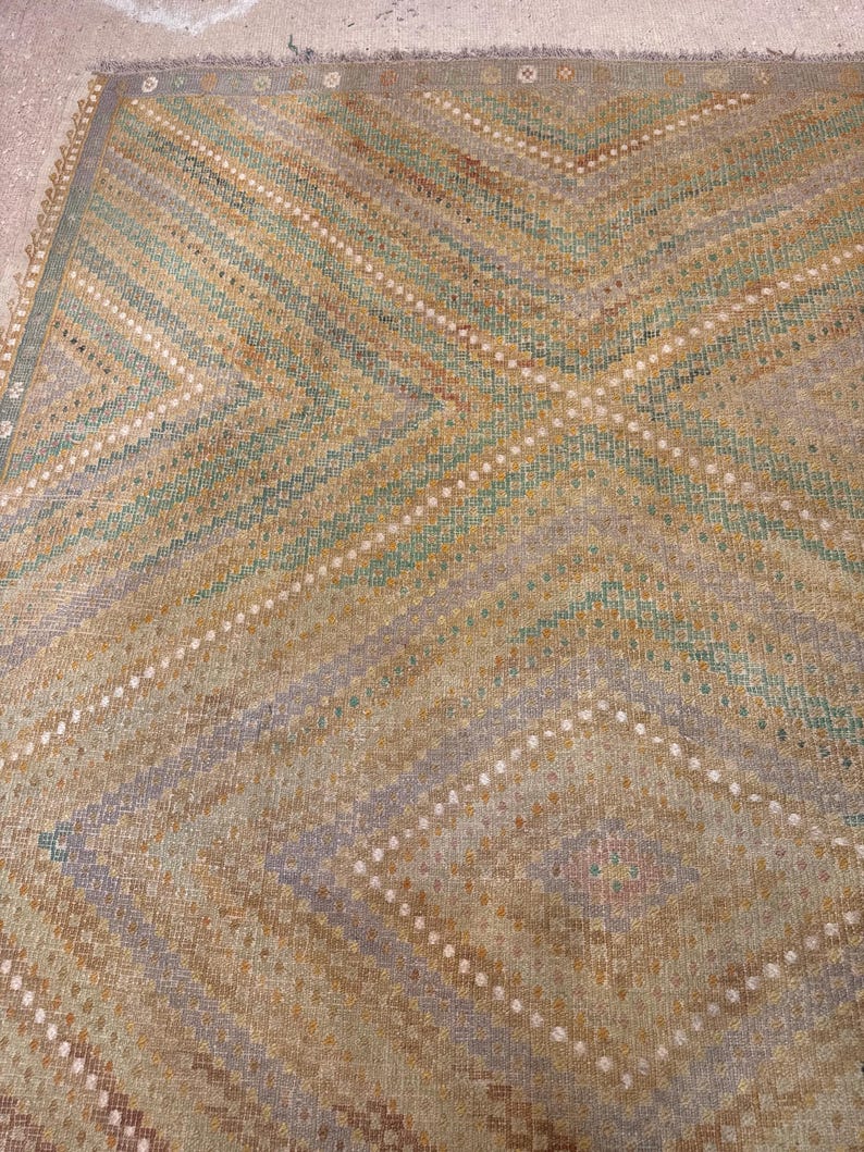 May include: A vintage rug with a geometric pattern. The rug features a diamond and chevron design in shades of tan, green, and gray. White dotted lines accent the pattern. The rug has a fringed edge and a worn appearance.