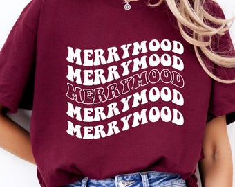May include: Burgundy t-shirt featuring the white text "MERRYMOOD" in a wavy design, repeated five times. The shirt is worn with blue denim jeans. The style is casual and relaxed.