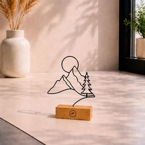 Puede incluir: Una escultura de alambre negro que representa una cordillera, un sol y un pino, montada sobre una base rectangular de madera clara. La base de madera presenta un pequeño logotipo circular con un símbolo de rayo centrado en la cara frontal.