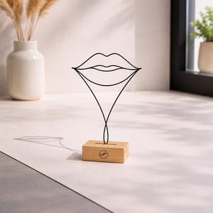 May include: A black wire sculpture shaped like a pair of lips, mounted on a rectangular light-colored wooden base. The base features a small circular logo centered on the front face. The wire structure forms a continuous outline of lips.