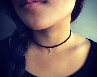 Puede incluir: Un collar tipo gargantilla de cordón negro con un pequeño dije de media luna en tono plateado centrado en el cordón. El collar se ajusta al cuello, ofreciendo una estética simple y limpia con un dije de luna metálico como punto focal.