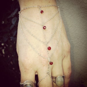 May include: A silver-toned hand chain bracelet featuring multiple delicate linked chains that drape across the back of the hand. The design includes four small, faceted red beads spaced along the central vertical chain, connecting to a ring loop.