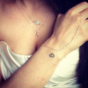 May include: A silver layered necklace featuring a small oval stone pendant and a wishbone charm, paired with a delicate silver hand chain bracelet that connects the wrist to a ring on the finger, worn with a white satin garment.