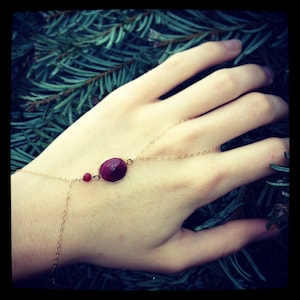 May include: A gold-toned hand chain featuring a delicate metal link chain, a small round red bead, and a larger oval faceted red gemstone set in a gold-coloured bezel. The jewellery piece connects from the wrist to the middle finger.