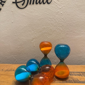 May include: A selection of coloured hourglasses in orange, blue, and teal. The sand timers are displayed on a wooden surface, with the word "Smile" visible in the background.