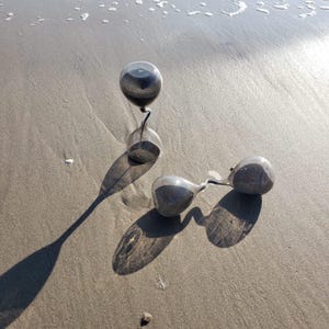 May include: Two silver hourglasses on a sandy beach. One hourglass is upright, casting a long shadow, while the other lies on its side. The sand is wet, and the ocean is visible in the background.