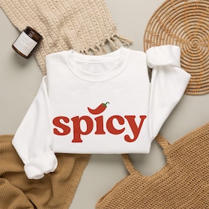 Op de afbeelding: Een wit sweatshirt met lange mouwen en een ronde hals, waarop het woord spicy in een vetgedrukt rood lettertype over het midden van de borst is gedrukt. Boven de letter i staat een kleine afbeelding van een rode chilipeper met een groene steel.