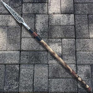 Puede incluir: Una lanza con una hoja plateada y un asta de madera. La hoja tiene un diseño intrincado. El asta es marrón y beige con detalles en negro y rojo. La lanza está sobre una superficie de ladrillos grises.