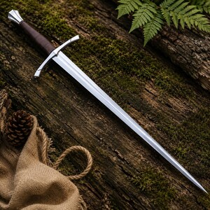 May include: A double-edged steel sword featuring a dark brown leather-wrapped grip, a polished silver crossguard, and a matching silver pommel. The blade is long, tapered, and pointed, reflecting light along its central ridge and sharp edges.