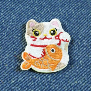 Peut inclure: Écusson brodé représentant un chat blanc avec des détails bruns et roses, tenant un poisson orange. Le chat a de grands yeux jaunes et un ruban rouge. L'écusson est sur un fond en denim bleu.