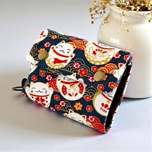 Puede incluir: Una pequeña cartera plegada con un estampado de gato Maneki Neko japonés. La cartera presenta un fondo azul marino con ilustraciones de gatos blancos, rojos y dorados y detalles florales. Tiene un cierre a presión y un pequeño anillo de metal.