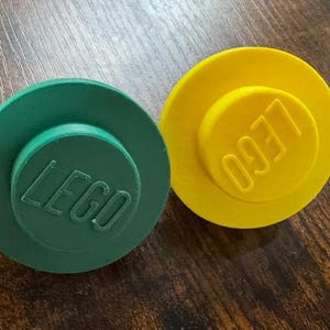 May include: Two circular plastic objects shaped like building block studs, one in dark green and one in bright yellow. Each piece features the embossed LEGO logo on the top surface, with a raised central circle and a flat, wide circular base.