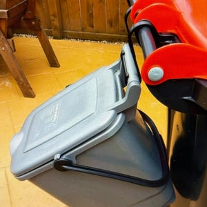 May include: A small grey plastic food waste bin with a hinged lid, suspended from the side of a larger black waste container with a bright orange lid. The grey bin features a black handle and a rectangular lid with printed recycling symbols.