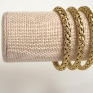 May include: Three gold-colored braided bracelets displayed on a beige fabric jewelry holder. The bracelets have a woven design and are circular in shape. The jewelry holder is cylindrical and has a light beige color.