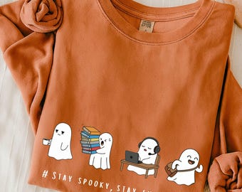 Op de afbeelding: Roestkleurige sweatshirt met een afbeelding van cartoongeesten in verschillende werksituaties. Een geest houdt een kopje koffie vast, een ander draagt boeken, een derde werkt op een laptop. Tekst: # STAY SPOOKY, STAY EMPLOYED #.