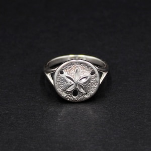 Könnte beinhalten: Ein silberner Ring mit einem Sanddollar-Motiv. Der Ring hat eine runde Oberfläche mit einem detaillierten Seesternmuster. Das Band ist glatt. Der Ring ist vor einem schwarzen Hintergrund platziert.