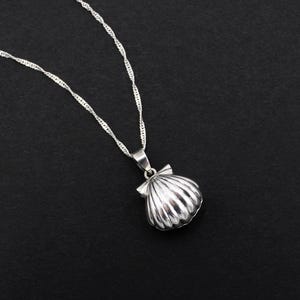 Op de afbeelding: Een zilveren ketting met een schelpenhanger op een zwarte achtergrond. De hanger heeft een gedetailleerd, driedimensionaal schelpenontwerp. De ketting is een fijne, gedraaide touwstijl.