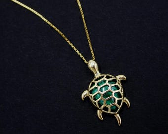 May include: A gold-toned necklace featuring a turtle-shaped pendant. The pendant has a polished gold frame with a green, patterned inlay within the shell section. The necklace includes a thin, delicate gold-toned box chain attached to the top.