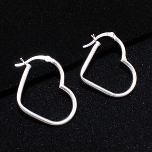 May include: A pair of silver-colored heart-shaped hoop earrings. The earrings are displayed on a dark, textured surface. The hoops are simple and elegant, with a classic design.