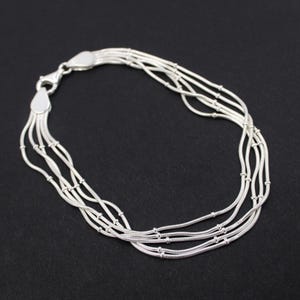 Op de afbeelding: Een zilveren armband met meerdere strengen, elk bestaande uit dunne, onderling verbonden schakels en kleine, ronde kralen. De armband heeft een veilige sluiting en ligt op een donkere achtergrond.