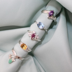May include: Five silver rings with various gemstone settings are displayed on a light blue fabric. The rings feature gemstones in shades of purple, blue, yellow, and red. One ring has a green teardrop-shaped stone.