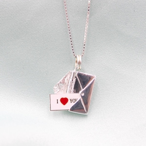 May include: A silver-colored pendant necklace featuring a small, open envelope charm. Inside the envelope is a tiny card with the words "I ❤️ YOU" and a red heart. The necklace has a delicate chain.