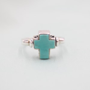 Puede incluir: Un anillo de plata con un diseño de cruz turquesa. La cruz es el punto focal, engastada en un marco cuadrado con elementos decorativos. El anillo tiene una banda sencilla.