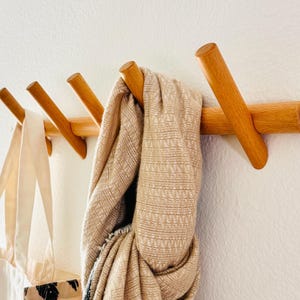 May include: Wooden coat rack with four angled pegs. A beige scarf and a tote bag hang from the pegs. The rack is made of light-colored wood and is mounted on a white wall. The scarf has a textured pattern.