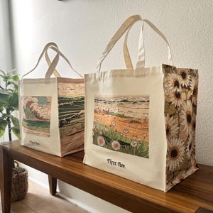 May include: Two canvas tote bags with cream handles and bodies. One bag features a coastal scene with a wave, the other a beach with daisies. The right side of one bag has a sunflower pattern. The text "Flynt Ave." is printed on the bottom of one bag.