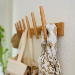 Op de afbeelding: Een houten wandkapstok met zes schuine haken. Een beige sjaal met wit patroon hangt aan de meest rechtse haak, terwijl een canvas tas en een bruine hoed aan de andere haken langs de houten lat zijn opgehangen.