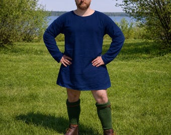 May include: A navy blue tunic with long sleeves and a rounded neckline. The tunic is paired with green knee-high socks and brown leather boots. The person is standing on a grassy field.