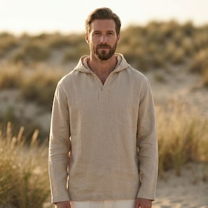 Op de afbeelding: Een beige linnen shirt met capuchon en lange mouwen. Het shirt heeft een V-hals opening en een relaxte pasvorm. Het model staat buiten voor een vage achtergrond van hoog gras en zand.